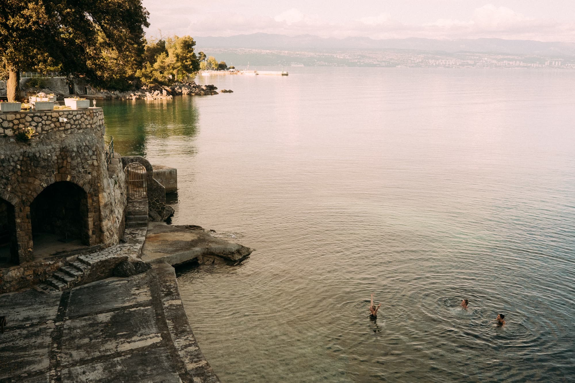 Swimmers in open water beside a stone wall, one with arms raised.